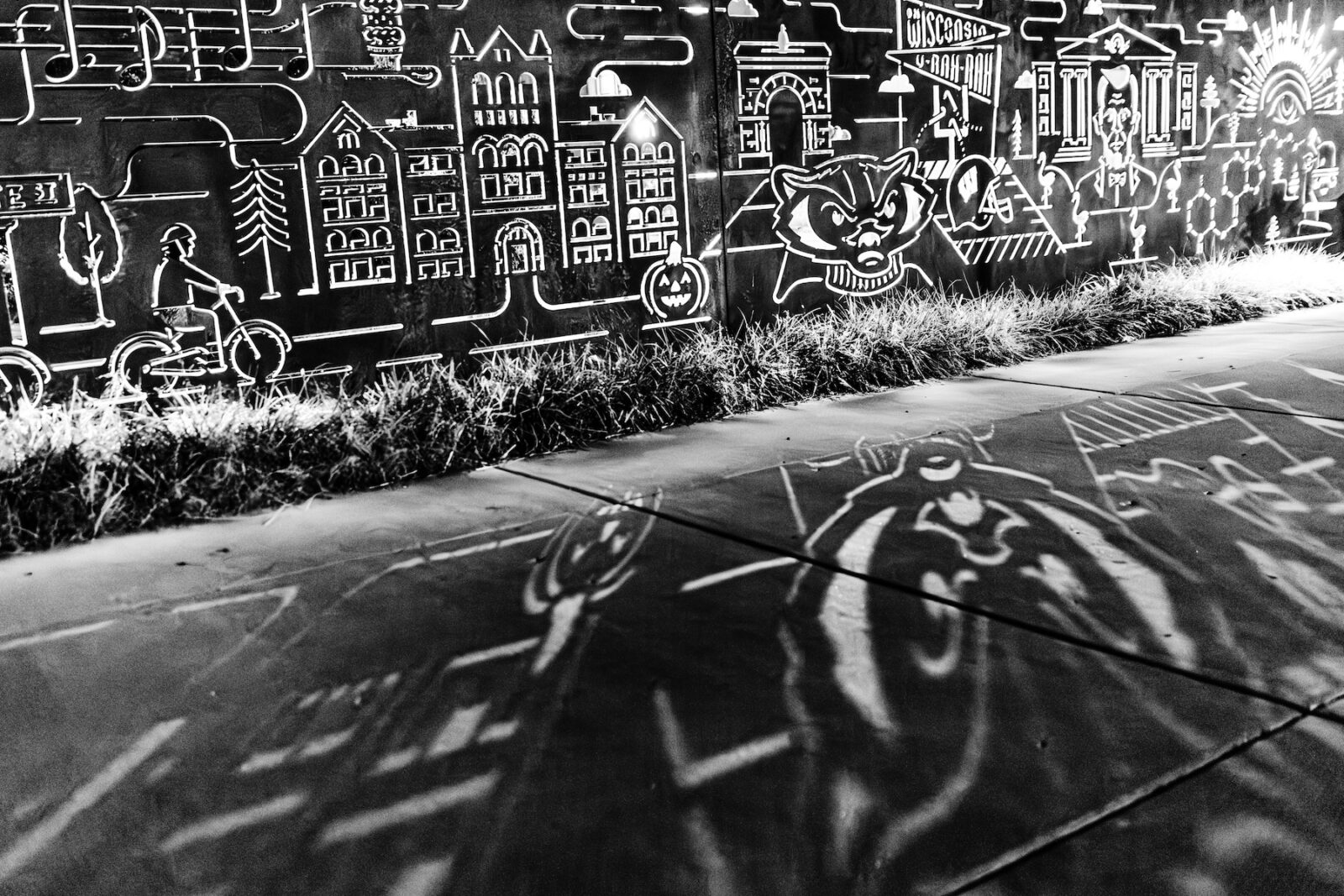 A shadow of Bucky Badger shines onto the sidewalk in Alumni Park at the University of Wisconsin-Madison during a cool autumn night on Sept. 18, 2023. Backlight in the background are iconic, cutout graphics in rusted steel of the park’s Badger Pride Wall. (Photo by Taylor Wolfram / UW–Madison)